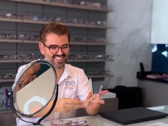 Atendente da ótica mostrando uma armação na mesa de atendimento, com um espelho redondo à frente